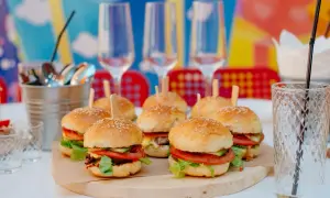 Mini hamburgery z pomidorami i sałatą na drewnianej desce, kieliszki i słomka w tle.