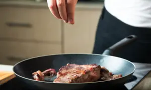 Ręka doprawia stek smażony na patelni.
