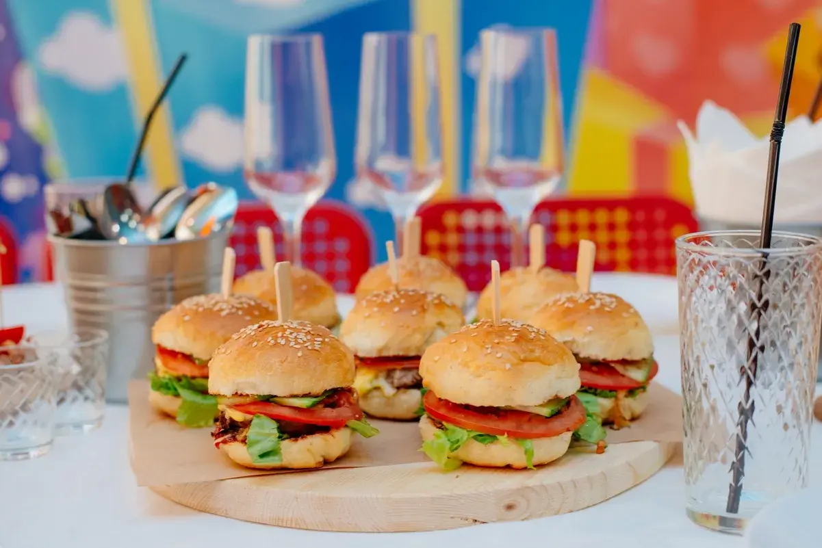 Mini hamburgery z pomidorami i sałatą na drewnianej desce, kieliszki i słomka w tle.