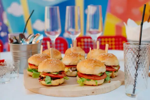 Mini hamburgery z pomidorami i sałatą na drewnianej desce, kieliszki i słomka w tle.